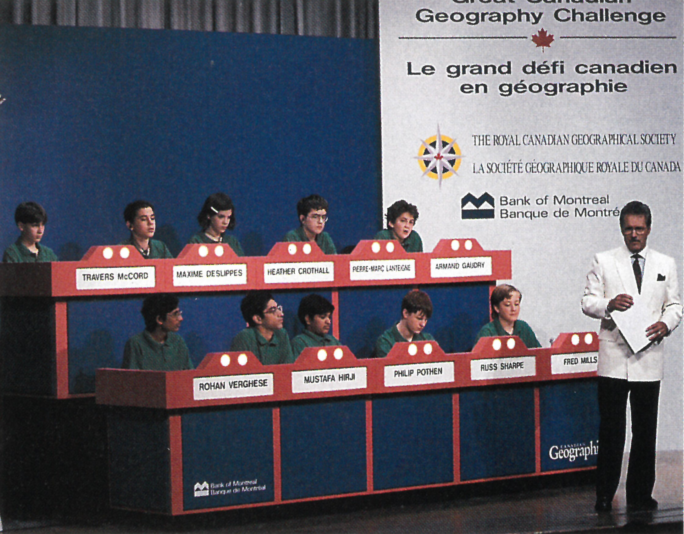 Alex Trebek, in a white blazer, stands next to two rows of quiz competitors