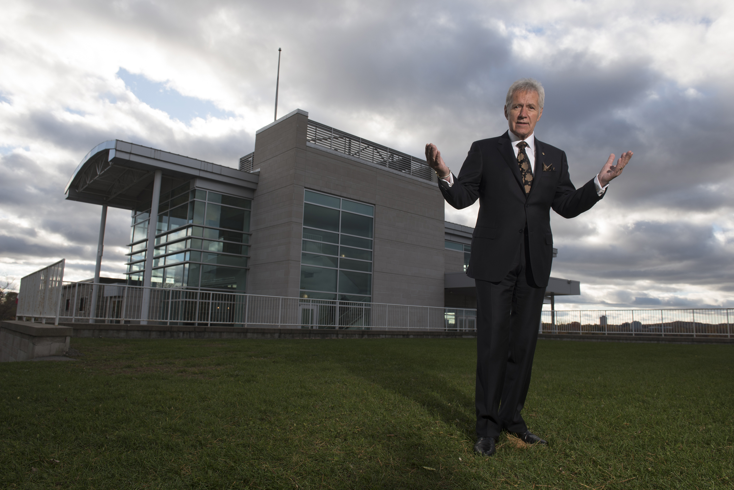Alex Trebek stands on grass with the Royal Canadian Geographical Society Headquarters behind him
