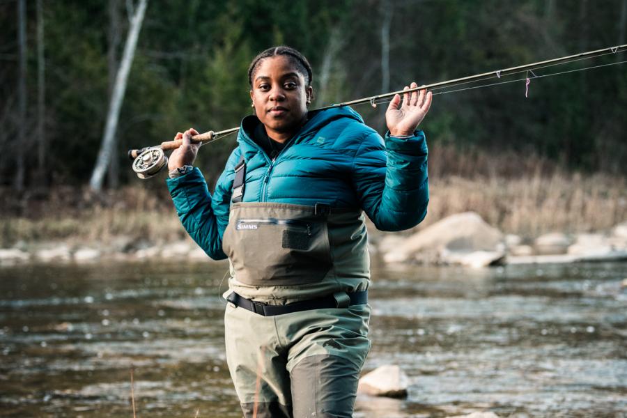 A woman dressed in fishing gear and a blue-green coat holds a fishing rod over her shoulders in a river
